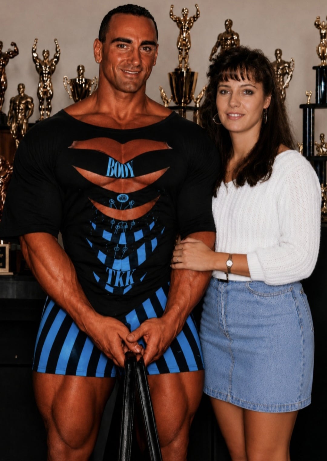 Muscular man in black and blue athletic wear poses with a smiling woman, trophies visible on shelves behind them.